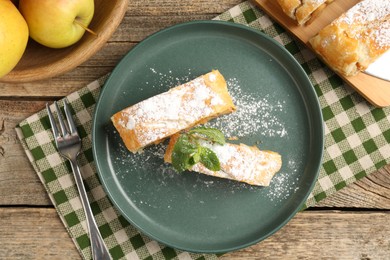 Photo of Pieces of tasty apple strudel with powdered sugar, mint, fruits and fork on wooden table, flat lay