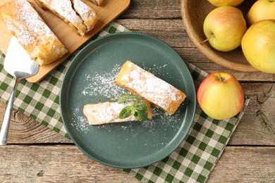 Photo of Pieces of tasty apple strudel with powdered sugar, mint, fruits and spatula on wooden table, flat lay