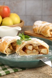 Photo of Pieces of tasty apple strudel with powdered sugar, mint and fork on wooden table, closeup