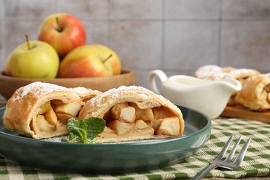 Photo of Pieces of tasty apple strudel with powdered sugar, mint and fork on table against grey tiled wall, closeup