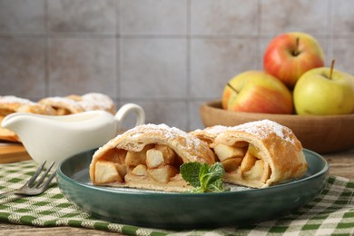 Photo of Pieces of tasty apple strudel with powdered sugar and mint on wooden table against grey tiled wall, closeup