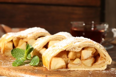 Photo of Pieces of tasty apple strudel with powdered sugar and mint on wooden board, closeup