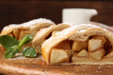 Photo of Pieces of tasty apple strudel with powdered sugar and mint on wooden board, closeup