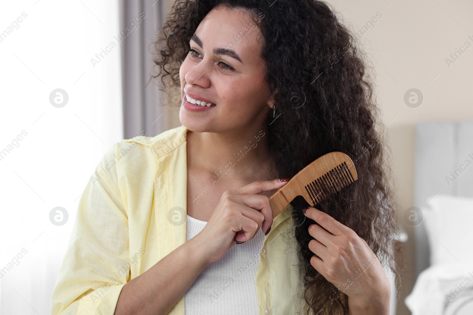 Photo of Smiling young woman brushing her curly hair with comb at home