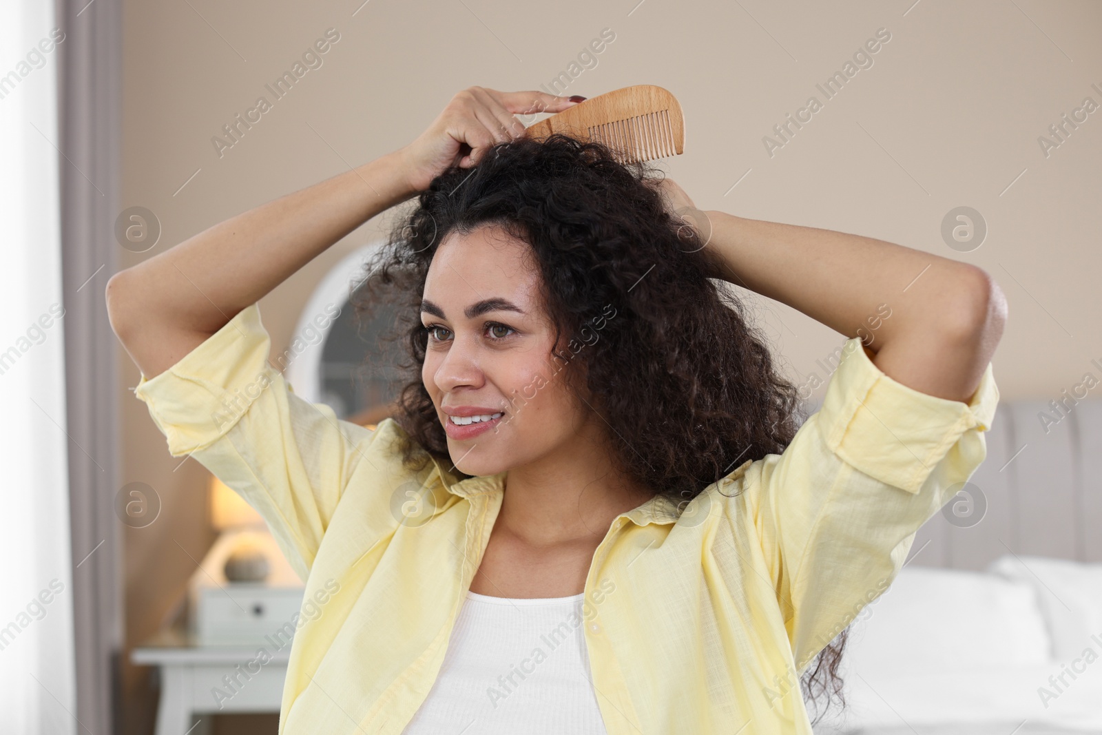 Photo of Smiling young woman brushing her curly hair with comb at home