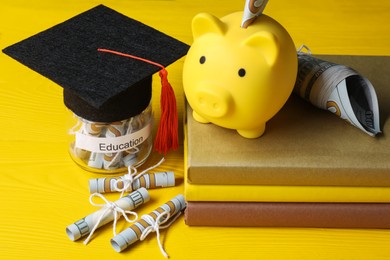 Photo of Piggy bank, graduate hat, books, dollar banknotes and glass jar with word Education on yellow wooden table, closeup. Tuition payment