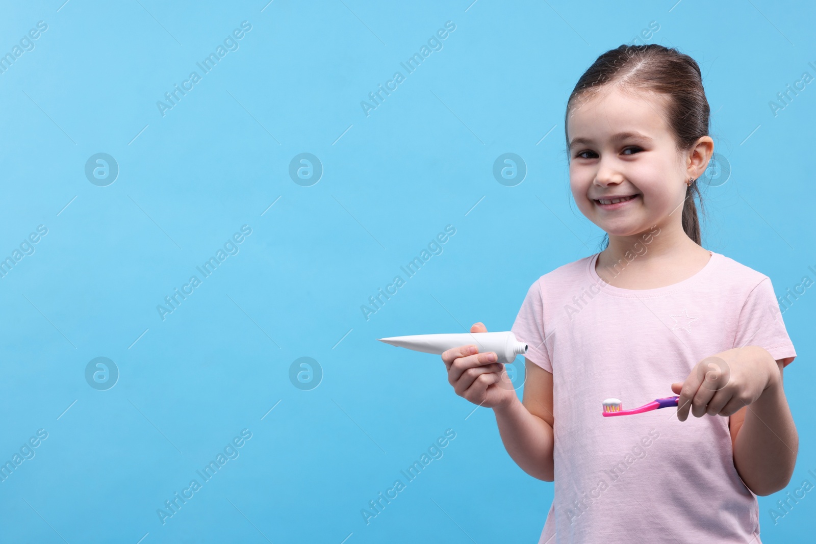 Photo of Cute girl with toothbrush and tube of toothpaste on light blue background, space for text