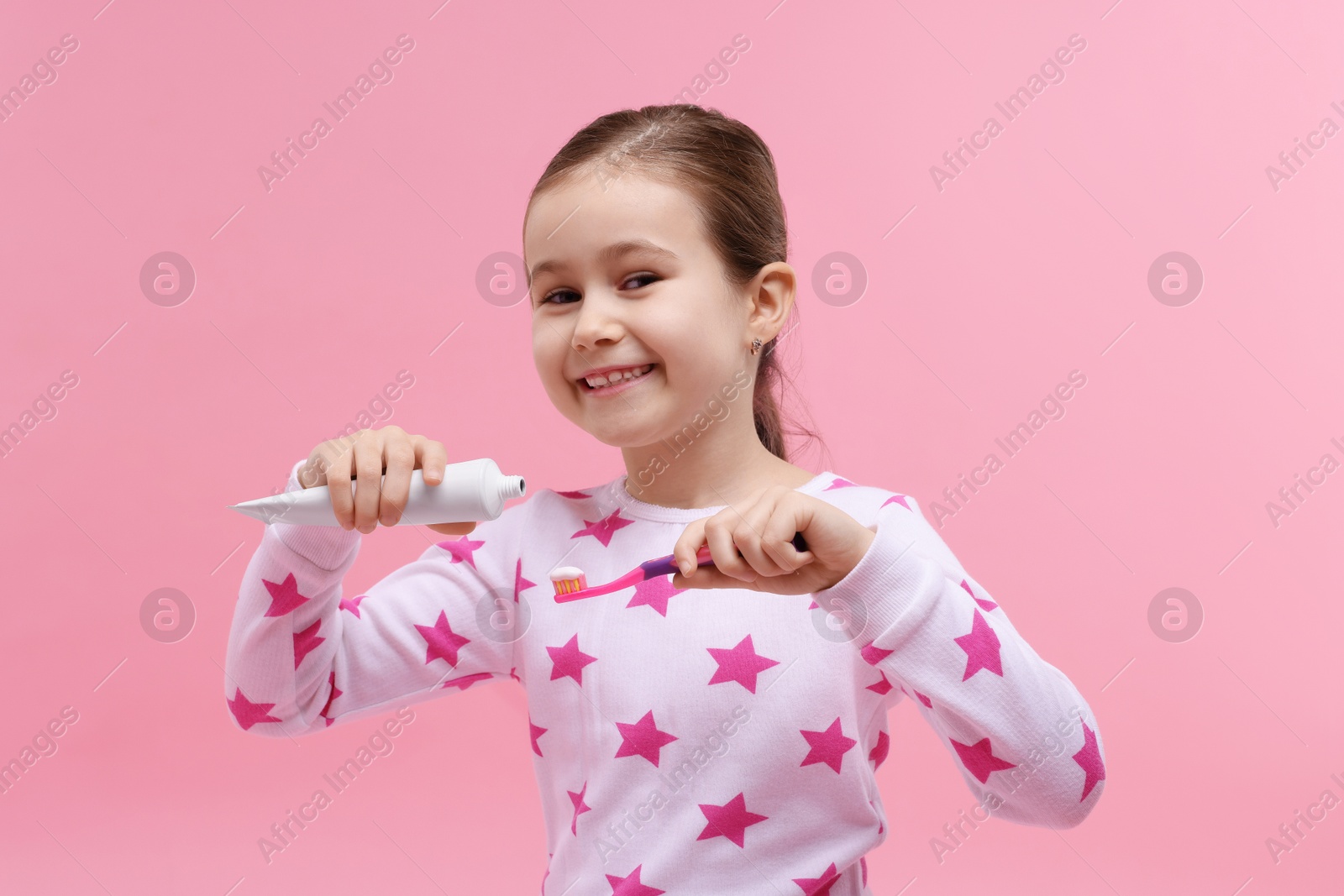 Photo of Cute girl applying toothpaste onto toothbrush on pink background