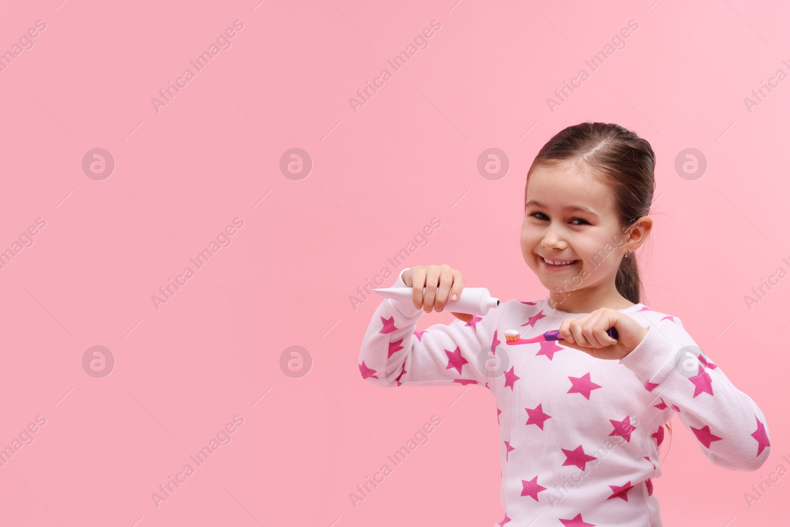 Photo of Cute girl applying toothpaste onto toothbrush on pink background, space for text