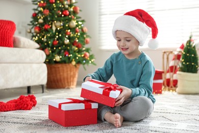 Photo of Happy little boy in Santa hat with Christmas gifts on floor at home