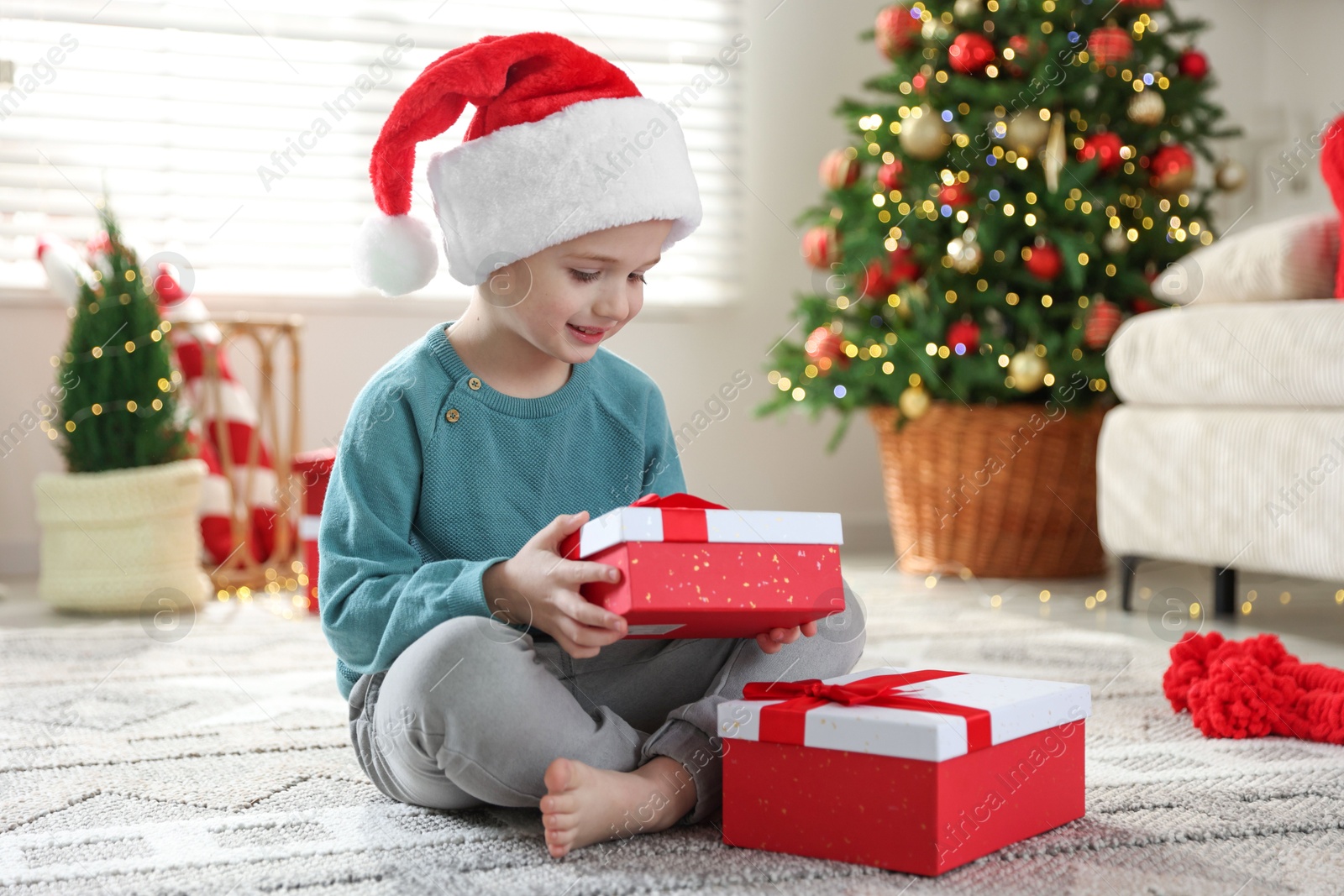 Photo of Happy little boy in Santa hat with Christmas gifts on floor at home