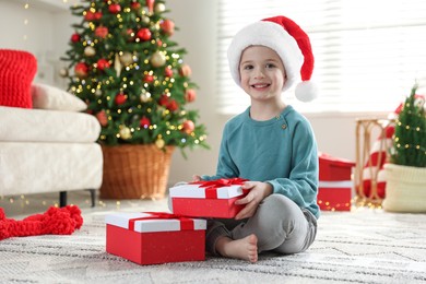 Photo of Happy little boy in Santa hat with Christmas gifts on floor at home