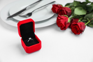 Photo of Engagement ring in box, rose flowers and tableware on white tablecloth, closeup. Romantic dinner