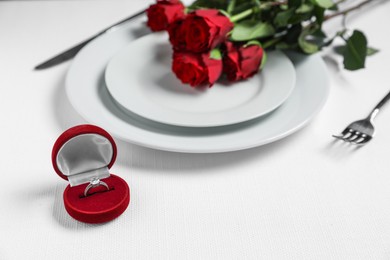 Engagement ring in box, flowers and tableware on white tablecloth, closeup. Table setting for romantic dinner