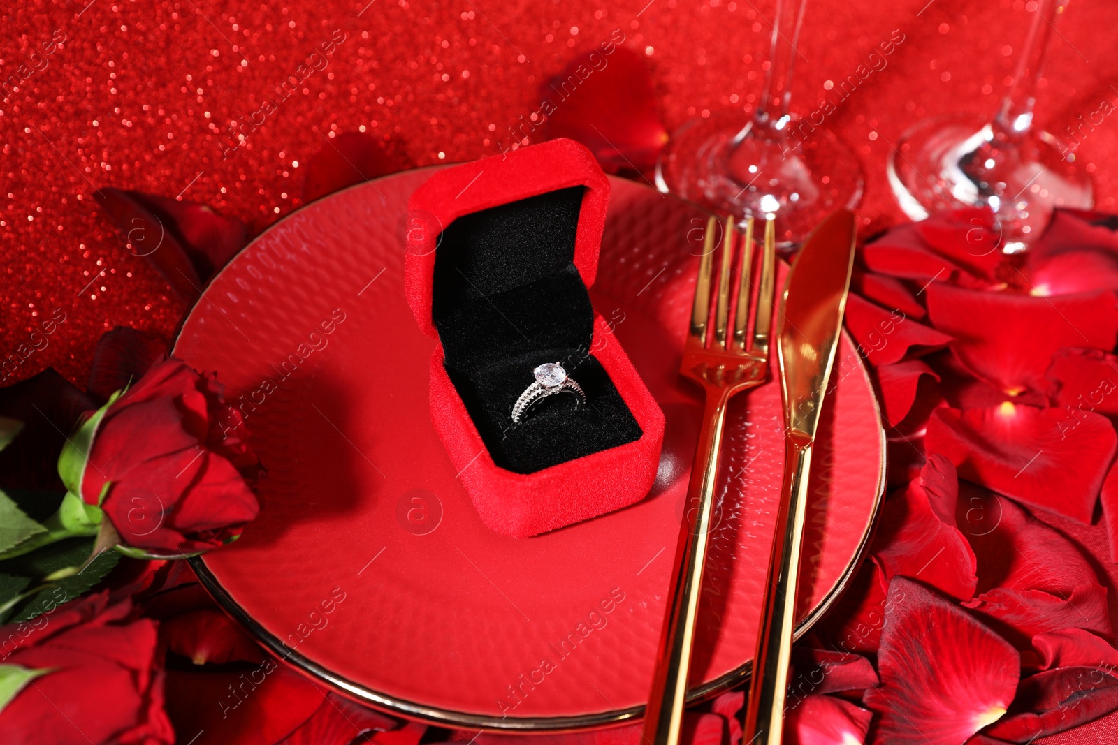 Photo of Engagement ring in box, tableware, rose flowers and petals on red glitter background, closeup. Table setting for romantic dinner