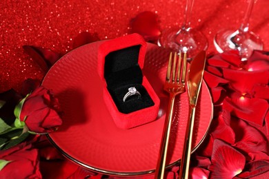 Photo of Engagement ring in box, tableware, rose flowers and petals on red glitter background, closeup. Table setting for romantic dinner