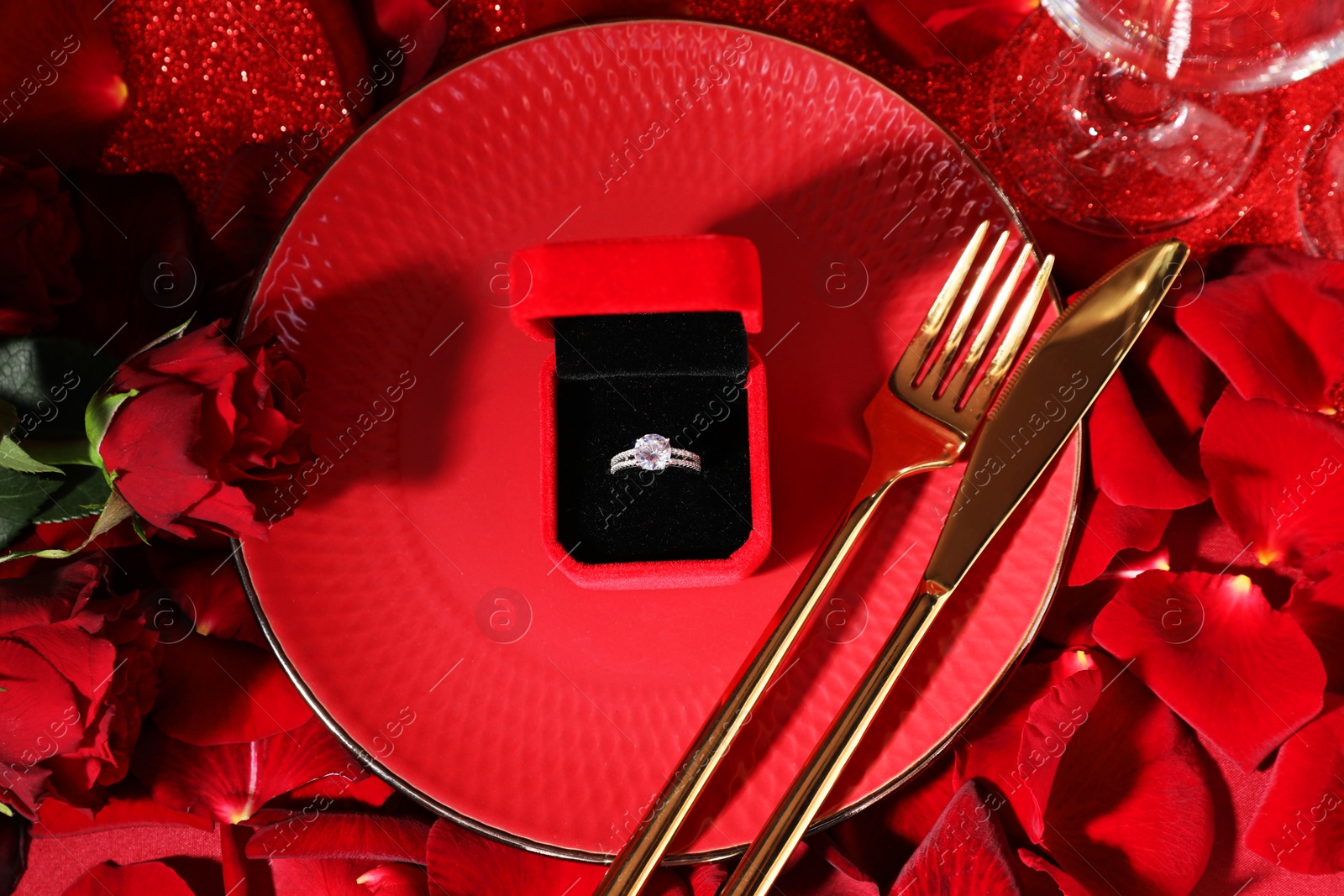 Photo of Engagement ring in box, tableware, rose flowers and petals on red glitter background, flat lay. Table setting for romantic dinner