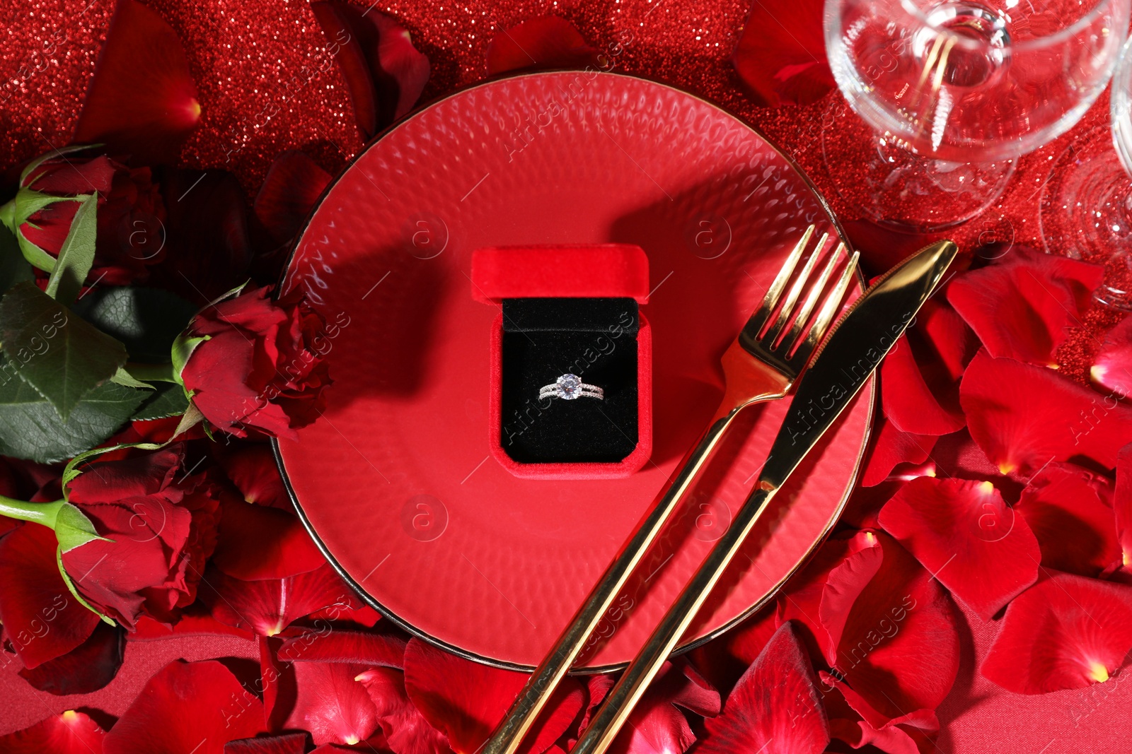 Photo of Engagement ring in box, tableware, rose flowers and petals on red glitter background, flat lay. Table setting for romantic dinner