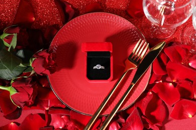 Engagement ring in box, tableware, rose flowers and petals on red glitter background, flat lay. Table setting for romantic dinner