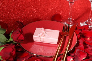 Gift box, plate, cutlery and roses on red glitter background, closeup. Beautiful table setting for romantic dinner