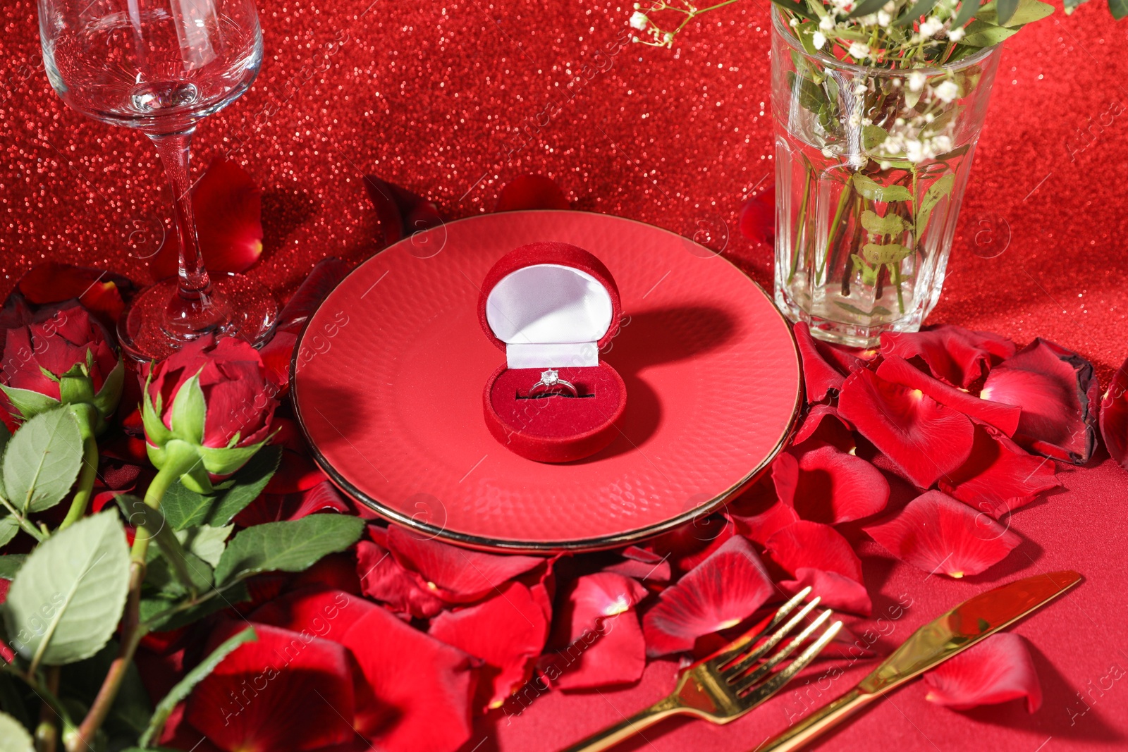 Photo of Engagement ring in box, tableware, rose flowers and petals on red glitter background. Table setting for romantic dinner