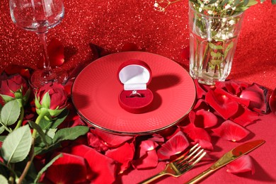 Photo of Engagement ring in box, tableware, rose flowers and petals on red glitter background. Table setting for romantic dinner