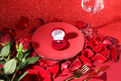 Engagement ring in box, tableware, rose flowers and petals on red glitter background. Table setting for romantic dinner