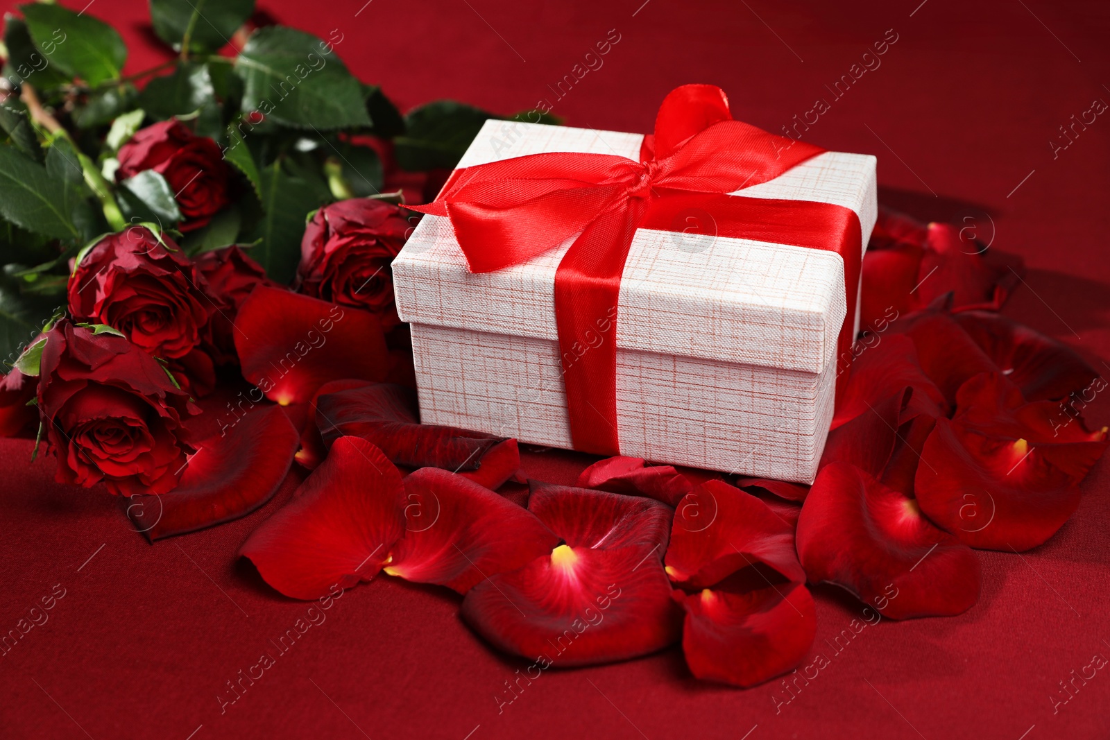 Photo of Beautiful gift box among rose flowers and petals on red background. Romantic present