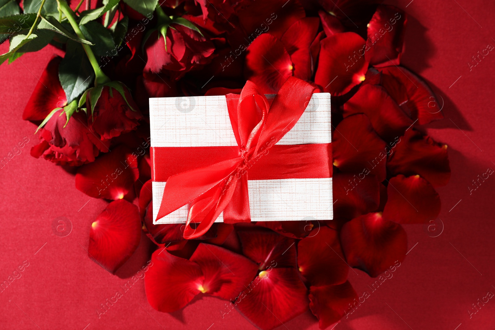 Photo of Beautiful gift box among rose flowers and petals on red background, flat lay. Romantic present