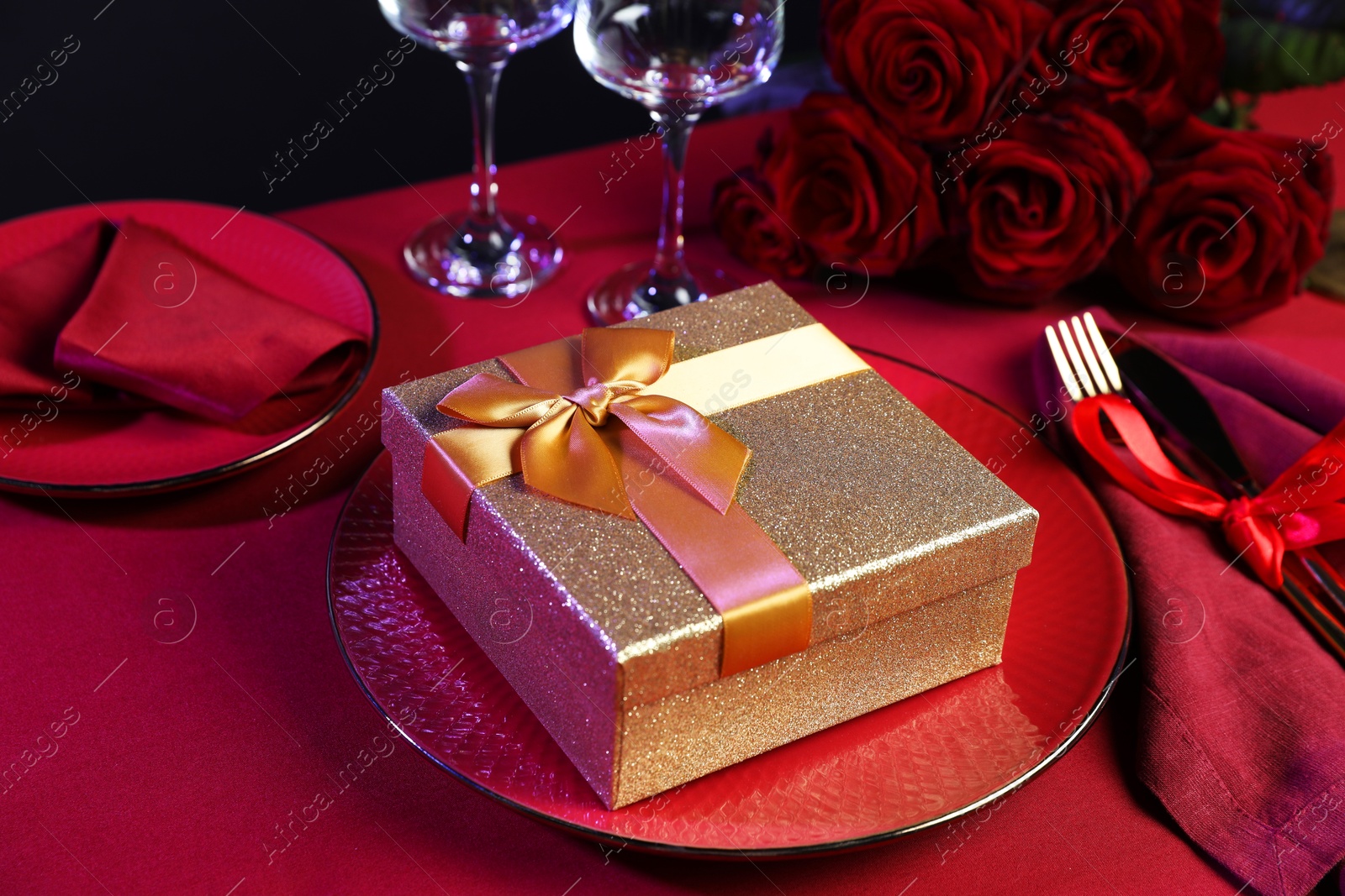 Photo of Gift box, tableware and roses on red table, closeup. Table setting for romantic dinner