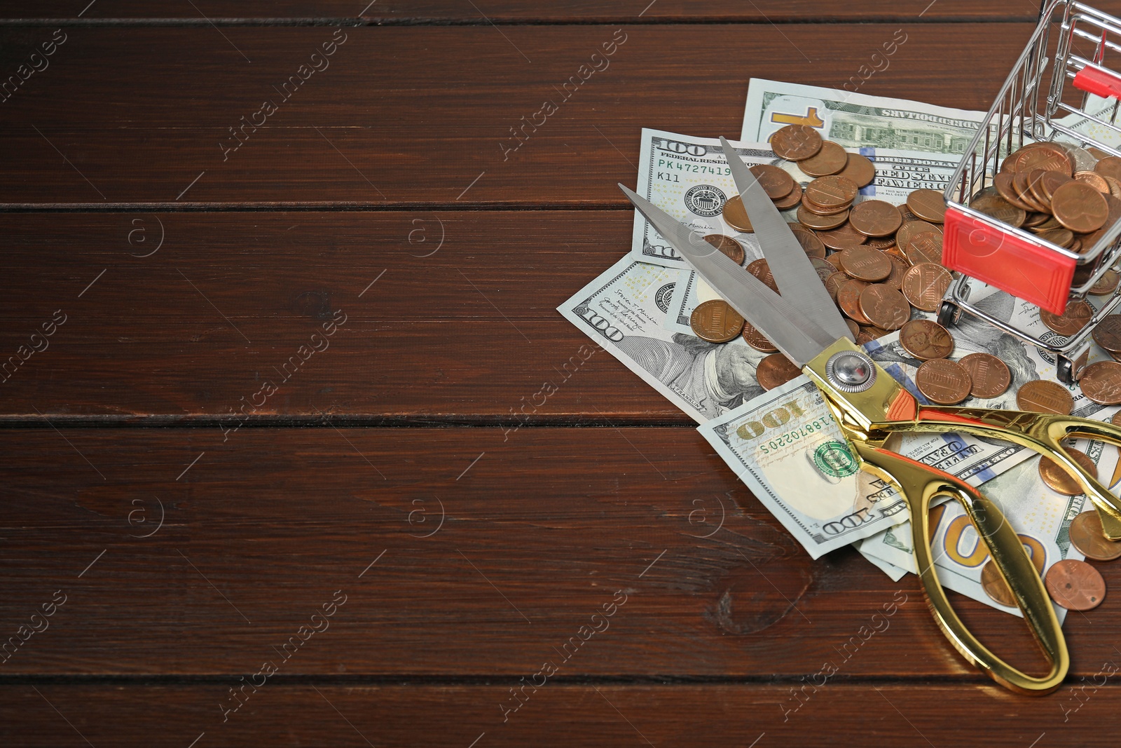 Photo of Dollar banknotes, scissors, coins and small shopping cart on wooden table, space for text