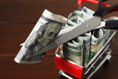 Photo of Dollar banknotes, scissors and small shopping cart on brown background, closeup