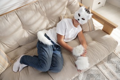 Photo of Quadrobics. Boy wearing cat mask, gloves and tail on sofa at home, above view