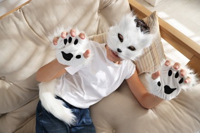 Photo of Quadrobics. Boy wearing cat mask, gloves and tail on sofa at home, above view