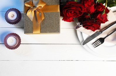 Beautiful table setting with romantic gift, flowers and burning candles on white wooden background, flat lay. Space for text