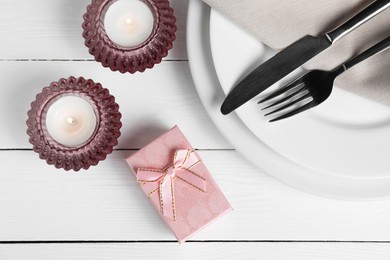 Beautiful table setting with romantic gift and burning candles on white wooden background, flat lay