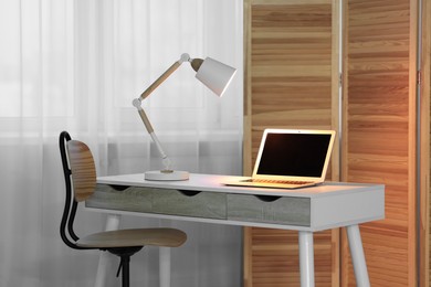 Photo of Comfortable workplace with laptop and lamp on table indoors