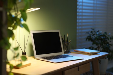 Photo of Comfortable workplace with laptop and lamp on wooden desk at home office in evening