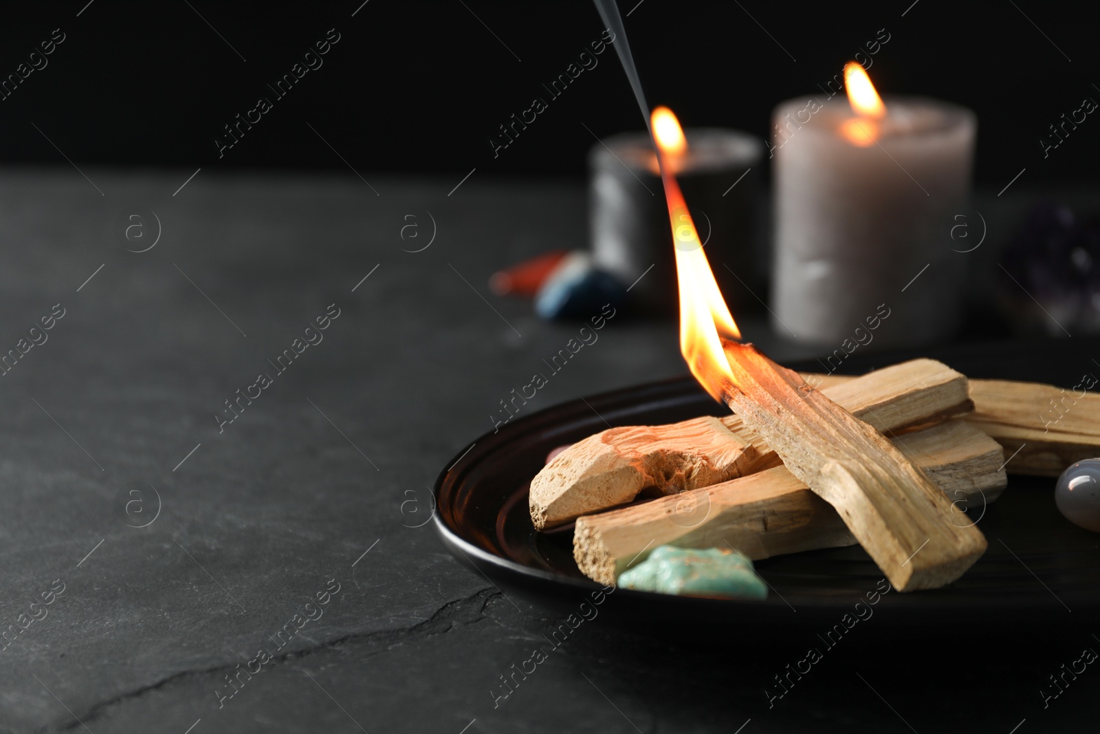 Photo of Burning palo santo stick, gemstones and candles on black table, closeup. Space for text