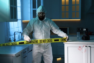 Photo of Forensic expert blocking way to crime scene with yellow tape indoors