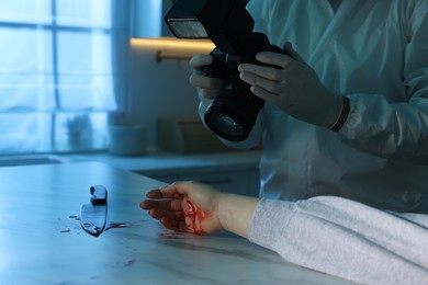 Photo of Forensic expert with camera, knife, blood and dead woman's body at countertop indoors, closeup