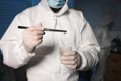 Photo of Forensic expert with tweezers collecting hair into container indoors, closeup