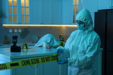 Photo of Forensic expert blocking way to crime scene and dead body with yellow tape indoors