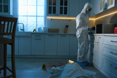 Photo of Forensic expert taking fingerprints with brush from countertop and dead body at crime scene indoors