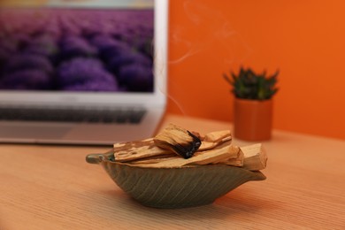 Photo of Palo santo sticks, laptop and houseplant on wooden table against orange wall