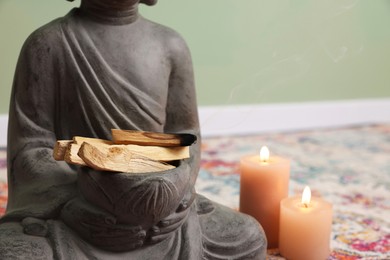 Photo of Buddha statue with palo santo sticks and burning candles indoors, closeup. Space for text