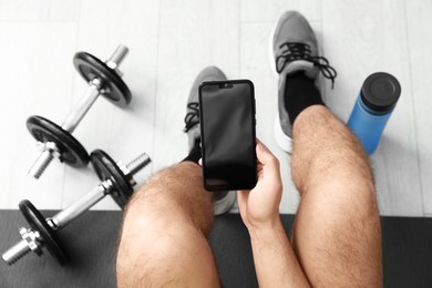 Photo of Man with smartphone, thermo bottle and barbells on floor indoors, above view