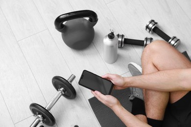 Photo of Man with smartphone and gym equipment on floor indoors, above view