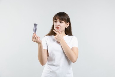 Pensive young woman with blister of contraceptive pills on light background