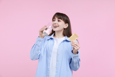 Young woman with contraceptive pills drinking water on pink background